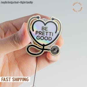 Pode incluir: Um porta-crachá com um design de estetoscópio em forma de coração. O design apresenta as palavras "BE PRETTI GOOD" em texto preto sobre um fundo perolado, com uma contorno dourado e preto. O porta-crachá é segurado em uma mão.