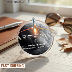 May include: A clear acrylic pin with the text "ARTEMIS II" over a depiction of the moon and a rocket launch. The pin also includes the text "We have liftoff" and the date and time of the launch.