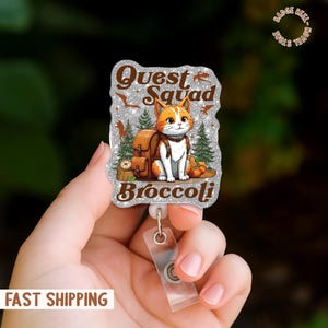 Könnte beinhalten: Ein Ausweis-Rollhalter mit glitzerndem silbernen Hintergrund, der eine Cartoon-Katze mit Rucksack, Kürbissen und Bäumen zeigt. Der Text lautet "Quest Squad Broccoli". Die Rolle hat einen transparenten Kunststoffriemen und einen Metallclip.