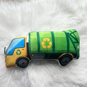 Pode incluir: Um brinquedo de pelúcia em forma de caminhão de lixo verde e amarelo. O caminhão tem um símbolo de reciclagem na lateral e uma cabine amarela.