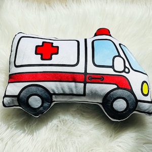 May include: A white plush ambulance pillow with a red cross, red stripe, and black outlines. The pillow has a blue windshield and yellow headlights.