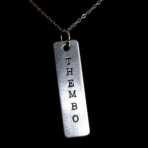 Puede incluir: Collar plateado con un colgante rectangular con la palabra "THEMBO" estampada verticalmente. La cadena del collar es delicada y cuelga sobre un fondo negro.