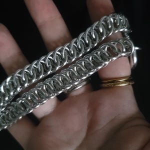 May include: Close-up of two silver chainmail bracelets. The bracelets are made of small, interlocking metal rings. The bracelets are resting on a hand, with a gold ring and silver rings visible.