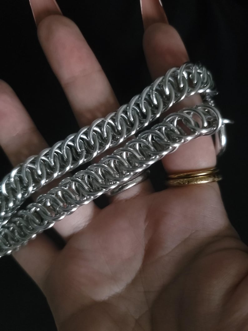 May include: Close-up of two silver chainmail bracelets. The bracelets are made of small, interlocking metal rings. The bracelets are resting on a hand, with a gold ring and silver rings visible.