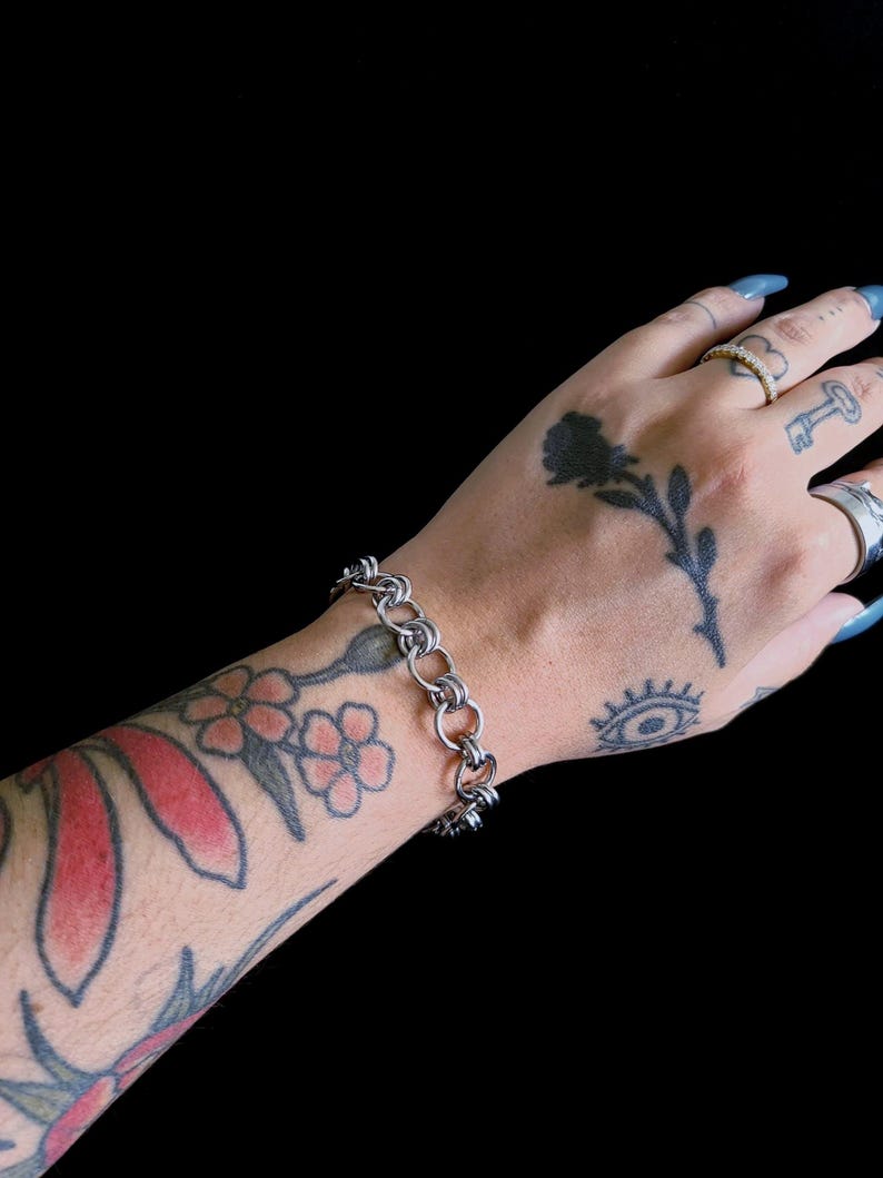 May include: A silver-toned chain bracelet featuring a series of interlocking circular links. The bracelet is constructed from polished metal rings connected in a repeating pattern, creating a flexible and continuous band around the wrist.