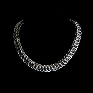 May include: A silver-colored chainmail necklace with a rounded design. The necklace is made of interlocking metal rings and is displayed against a black background. The chain is a medium thickness.