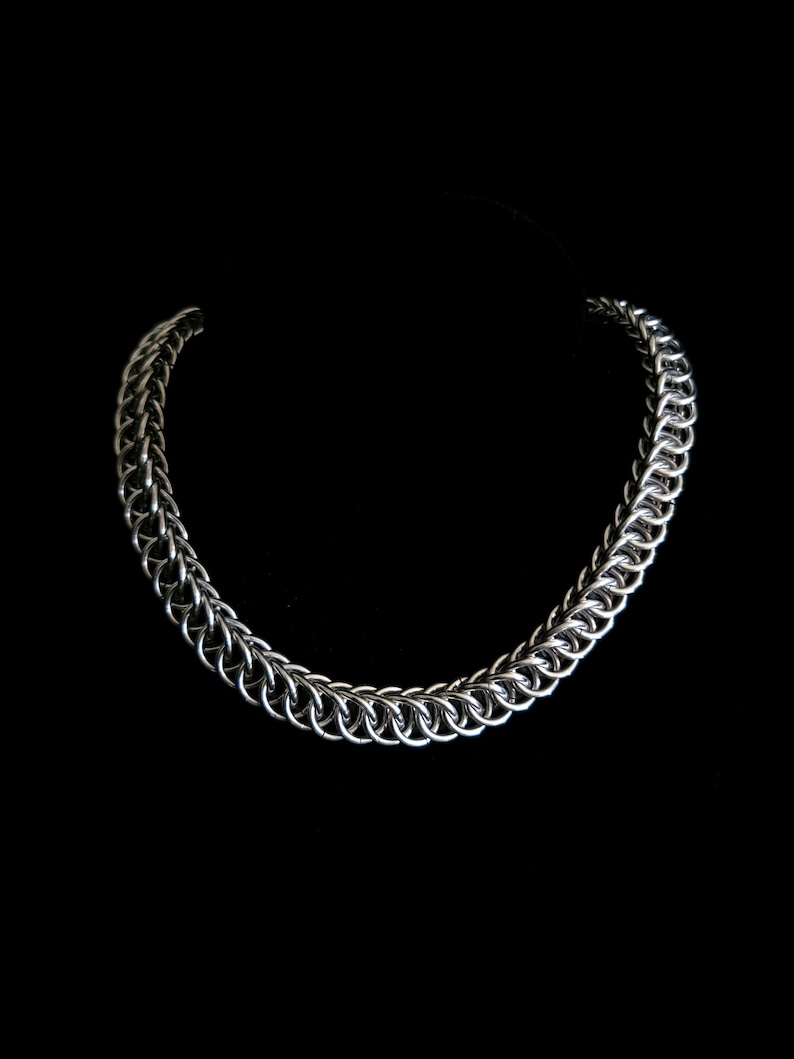 May include: A silver-colored chainmail necklace with a rounded design. The necklace is made of interlocking metal rings and is displayed against a black background. The chain is a medium thickness.