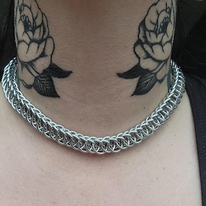 May include: A silver chainmail necklace with a close-knit design. The necklace is worn around the neck. The background includes black rose tattoos on the neck and a black tank top.