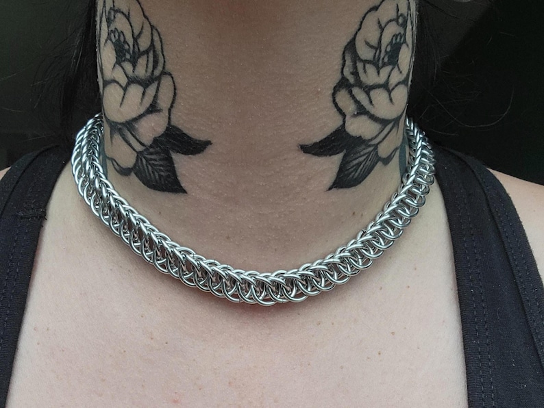 May include: A silver chainmail necklace with a close-knit design. The necklace is worn around the neck. The background includes black rose tattoos on the neck and a black tank top.