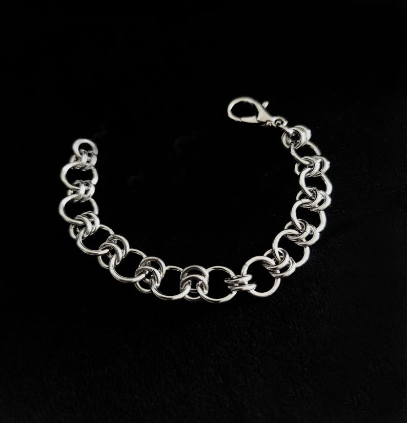 May include: A silver-toned chainmail bracelet constructed from a series of interlocking circular metal rings. The bracelet features a polished finish and is secured with a lobster claw clasp at one end, forming a continuous, flexible circular link design.