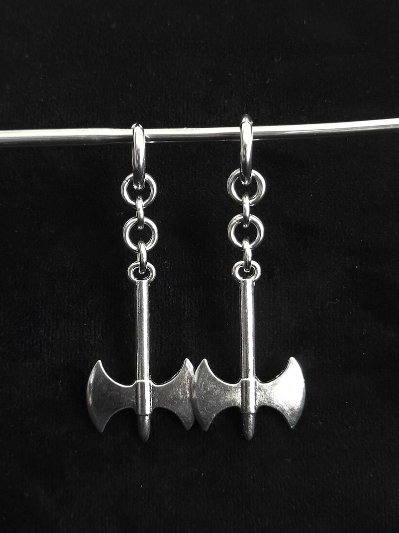 May include: Silver-toned axe earrings. Each earring features a small axe pendant suspended from a series of linked rings and a hoop. The axes have a double-bladed design and are displayed against a black background.