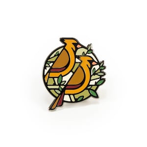 May include: A gold and orange enamel pin featuring two stylized birds in a circular design with green leaves. The birds are facing each other and are surrounded by a circle of green leaves.