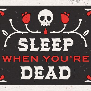 May include: A rectangular black sign with white text that reads "SLEEP WHEN YOU'RE DEAD" in a bold, vintage font. The design includes a skull, red roses, and decorative flourishes.