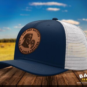 May include: Navy blue trucker hat with a white mesh back and a curved brim. The hat features a brown leather patch with the text "EAT HER SHITTER LIKE AN APPLE FRITTER" and a Grim Reaper graphic. The hat is made by Barton Trading Co.