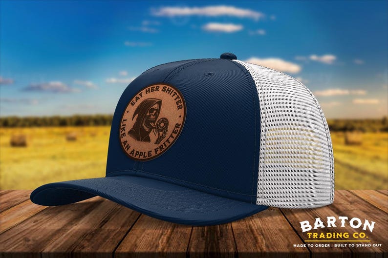 May include: Navy blue trucker hat with a white mesh back and a curved brim. The hat features a brown leather patch with the text "EAT HER SHITTER LIKE AN APPLE FRITTER" and a Grim Reaper graphic. The hat is made by Barton Trading Co.