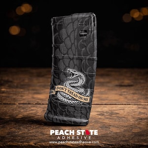 May include: A black magazine with a textured pattern and a graphic of a coiled snake with the text "DON'T TREAD ON ME" on a banner. The magazine is on a wooden surface.
