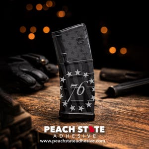 May include: Black magazine with a white star and stripe design, featuring the number "76". The magazine is angled on a wooden surface. The text "PEACH STATE ADHESIVE" and website address are visible at the bottom.