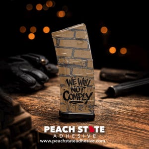 May include: A clear magazine featuring a brick wall design and the words "WE WILL NOT COMPLY" in black graffiti-style lettering. The magazine has a black base and is set on a wooden surface. The Peach State Adhesive logo is at the bottom.