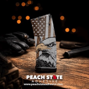 Puede incluir: Un cargador con un diseño patriótico que presenta una bandera estadounidense y una ilustración detallada de un águila. El diseño incorpora colores marrón, blanco y negro. El texto "PEACH STATE ADHESIVE" y la dirección del sitio web son visibles en la parte inferior.