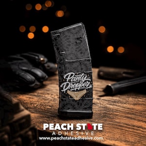 Puede incluir: Un cargador negro con un diseño decorativo y las palabras "Panty Dropper" en escritura blanca. Un gráfico dorado de ropa interior está debajo del texto. Las palabras "PEACH STATE ADHESIVE" están en la parte inferior.