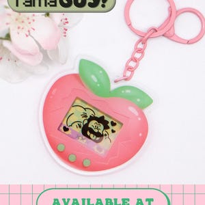 May include: A pink peach-shaped acrylic keychain featuring a digital screen graphic of a cartoon character with a mustache and hearts. The keychain includes a pink metal ring and chain. The text TamaGUY! appears at the top, and AVAILABLE AT ETSY.COM/SHOP/DREWDRAWSPINUPS is at the bottom.