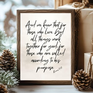 Op de afbeelding: Een rechthoekig ingelijst wandkunstwerk met een witte achtergrond en zwarte cursieve tekst die luidt: And we know that for those who love God all things work together for good, for those who are called according to his purpose Romans 8:28.