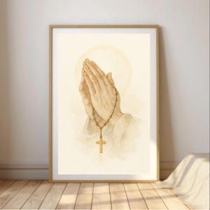 Puede incluir: Una impresión en acuarela enmarcada que representa manos juntas en oración, con un rosario y una cruz dorada. La obra de arte tiene una paleta de colores suaves y cálidos y un efecto de halo detrás de las manos. El marco es de color marrón claro.