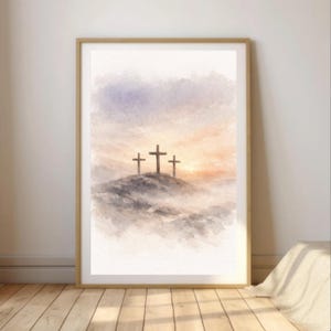 Puede incluir: Una impresión en acuarela enmarcada representa tres cruces sobre una colina, contra un cielo suave y pastel. La obra de arte presenta una paleta de colores apagados de azules, naranjas y grises, creando una escena serena. El marco es de color madera clara.