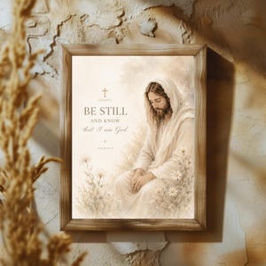 May include: A framed wall art print featuring a beige and cream watercolor illustration of a figure with long hair and a robe sitting among flowers. The text reads: BE STILL AND KNOW that I am God. PSALM 46:10, with a small cross symbol above.