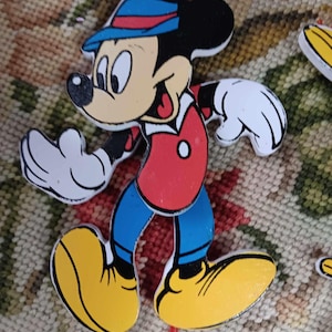 May include: A wooden Mickey Mouse character with a red and blue hat, red shirt, blue pants, and yellow shoes. The character is painted in a cartoon style and has a friendly expression.