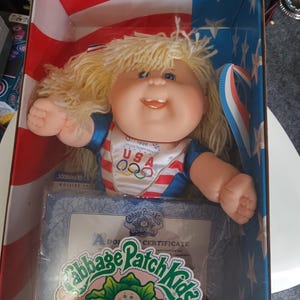 May include: A Cabbage Patch Kids OlympiKids Special Edition doll in its original box. The doll has blonde hair and is wearing a red, white, and blue outfit with the USA Olympic rings logo. The box features an American flag design.