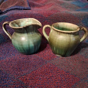 May include: Two ceramic pitchers with handles. One is teal with a ruffled rim, the other is olive green with two handles. Both have a glossy finish and sit on a red and blue textured surface. The pitchers are likely for serving.