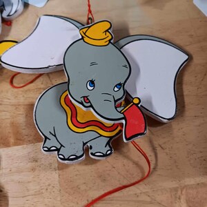 May include: A gray and white wooden toy elephant with a yellow hat and red and yellow trim. The elephant is attached to a red string with a pink bead at the end.