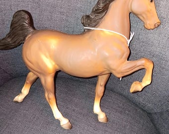 May include: A brown plastic horse figurine with a dark brown mane and tail. The horse is posed with one front leg raised. A white string is around the horse's neck. The horse is on a gray surface.