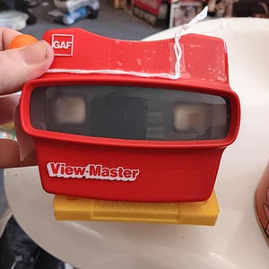 May include: A vintage red View-Master viewer with the GAF logo and the words "View-Master" in white. The viewer is held over a yellow slide holder. The viewer has two eye pieces and a white string attached to the top.