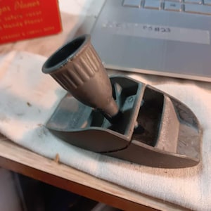 May include: A gray, vintage-style hand plane with a dark gray handle and a rectangular base. The plane appears to be made of metal and is resting on a white cloth. The handle has vertical ridges.