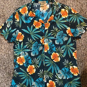 May include: A short-sleeved button-down shirt with a tropical floral pattern. The shirt features a dark teal background with orange and white flowers, and blue and green leaves. The brand name "notations" is visible on the tag.