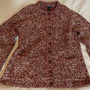 May include: A button-down, long-sleeved cardigan sweater in a speckled burgundy and cream knit. The sweater has a collared neckline, two front pockets, and a series of brown buttons down the front. The knitwear is a medium weight.