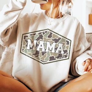 May include: A cream-colored sweatshirt with a camouflage design in a hexagonal shape, featuring the word "MAMA" in white block letters. The sweatshirt is worn with denim shorts and a black mug.