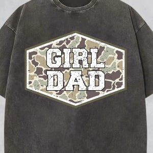 May include: Dark gray t-shirt with a distressed look. The shirt features a camouflage design within a white-outlined hexagon, with the words "GIRL DAD" in bold, white letters. The shirt has short sleeves and a crew neck.