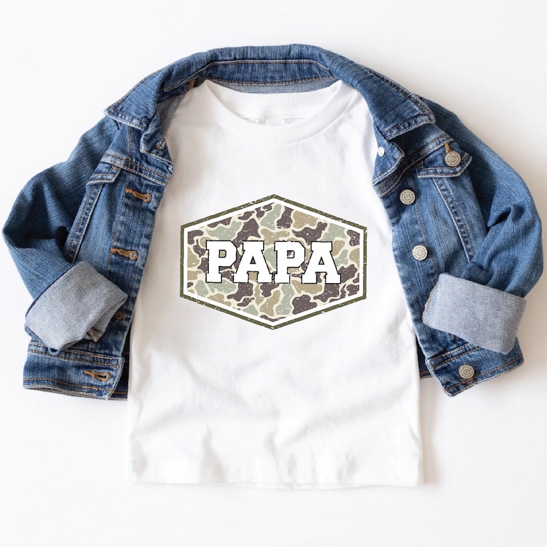 May include: White t-shirt with a camouflage design in a hexagonal shape, featuring the word "PAPA" in a bold, distressed font. The shirt is paired with a blue denim jacket.