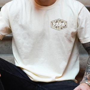 May include: Cream-colored t-shirt with a camouflage-patterned "PAPA" design in a hexagonal shape on the chest. The shirt is made of a soft material and has short sleeves. The design is in shades of green, brown, and tan.