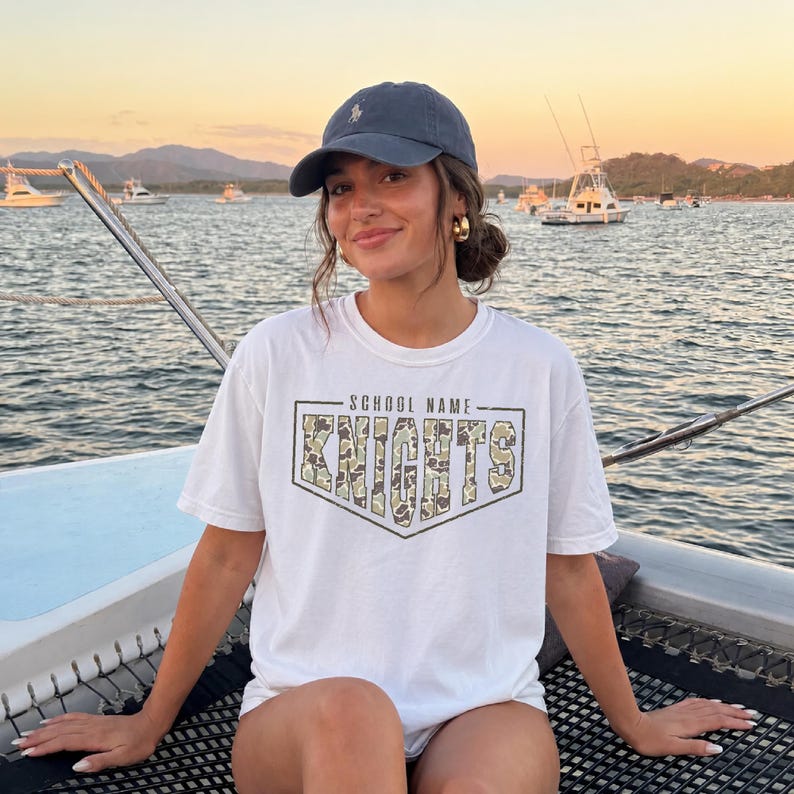 May include: A white short-sleeve t-shirt featuring a graphic design with the text SCHOOL NAME above the word KNIGHTS in a camouflage pattern. The shirt is paired with a dark navy blue baseball cap with a small embroidered logo on the front.