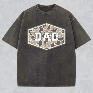 May include: Dark gray t-shirt with a distressed look. The shirt features a camouflage design in a hexagonal shape, with the word "DAD" in white, block letters. The shirt has short sleeves and a crew neck.