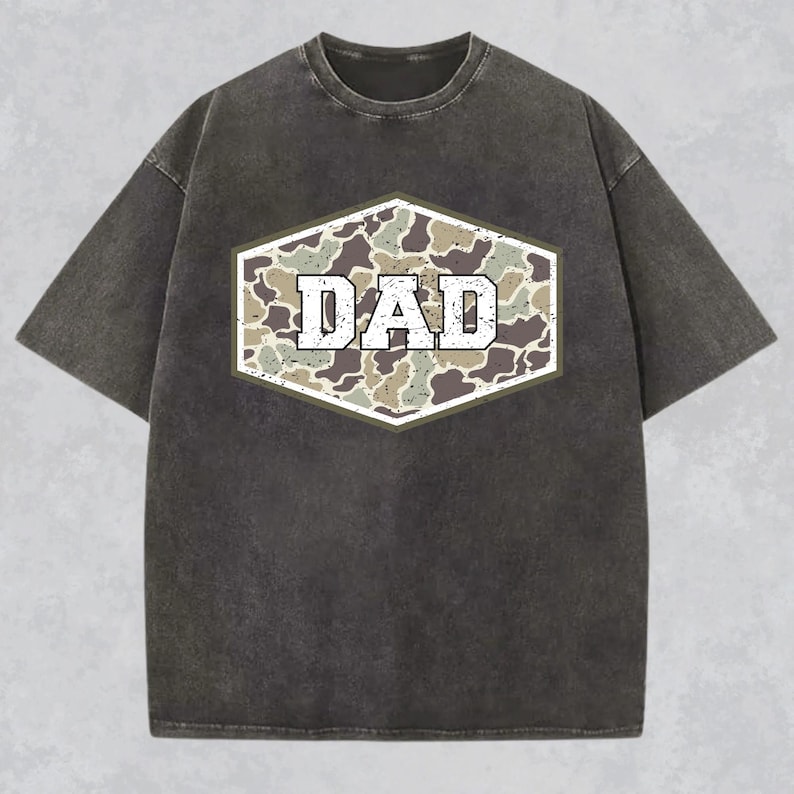 May include: Dark gray t-shirt with a distressed look. The shirt features a camouflage design in a hexagonal shape, with the word "DAD" in white, block letters. The shirt has short sleeves and a crew neck.