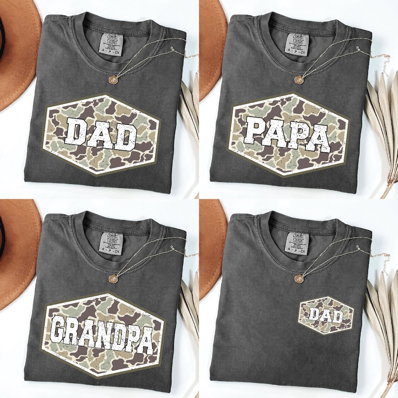 May include: Four dark gray t-shirts with the words "DAD", "PAPA", "GRANDPA", and "DAD" printed in white block letters inside a camouflage hexagon. Each shirt has a crew neck and a small gold necklace.