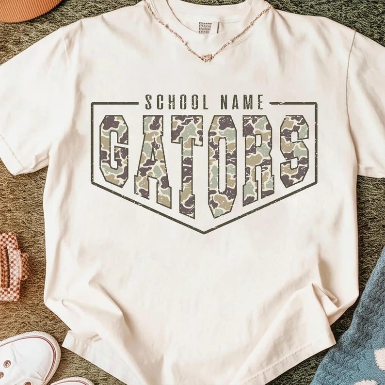 May include: A cream-colored short-sleeve t-shirt featuring a graphic design with the text "SCHOOL NAME" above the word "GATORS" in a large, block-letter font. The letters are filled with a green, brown, and tan camouflage pattern.