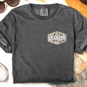 May include: Dark gray Comfort Colors t-shirt with a small chest pocket featuring the word "GRANDPA" in white lettering over a camouflage pattern. The shirt has a crew neck and short sleeves. A small tag is visible near the collar.