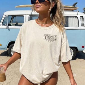 May include: A cream-colored short-sleeve t-shirt featuring a graphic on the left chest. The graphic is a shield-shaped outline containing the word TIGERS in a leopard print pattern with the text SCHOOL NAME printed above it in black ink.