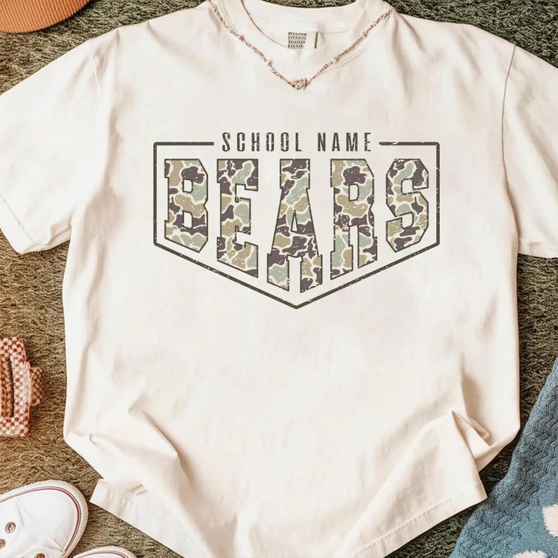 May include: A cream-colored short-sleeve t-shirt featuring a dark-outlined shield graphic. Inside the shield, the text "SCHOOL NAME" appears above the word "BEARS" in large block letters filled with a green, brown, and tan camouflage pattern.